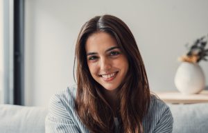 A young woman with long brown hair sits indoors, smiling at the camera. There is a white vase with dried flowers in the blurred background.